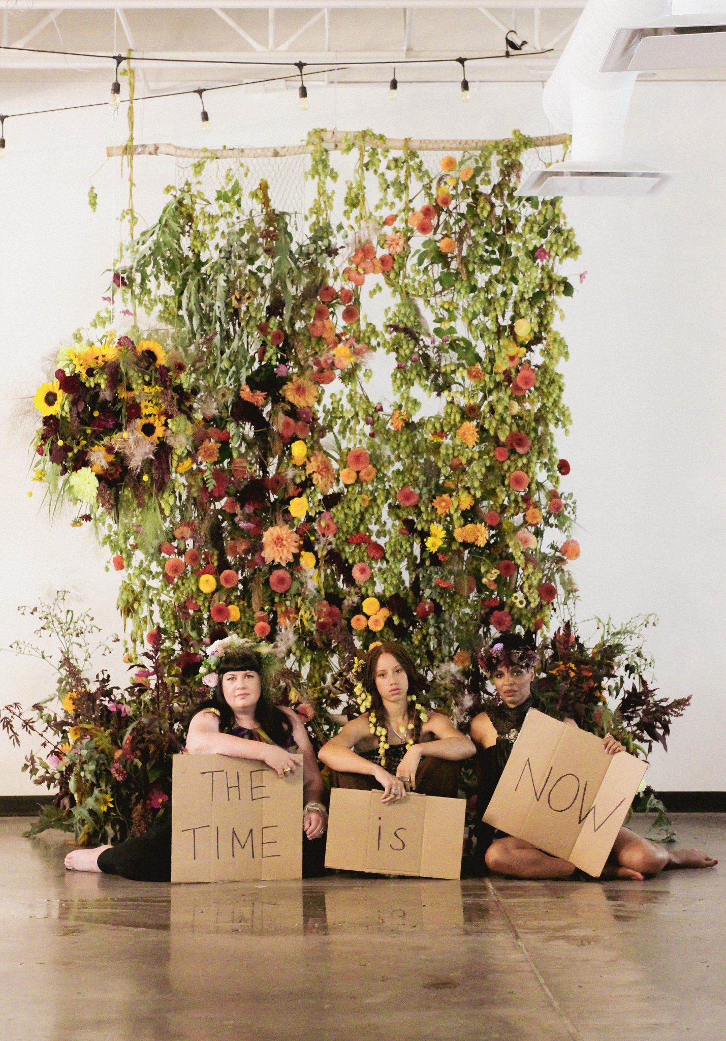 Three women holding cardboard signs that read 'The Time is Now' in front of a floral installation.