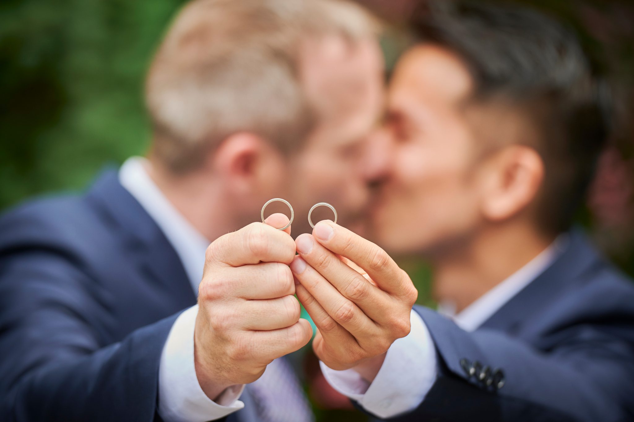 Same sex wedding couple hold up their wedding rings while kissing. 