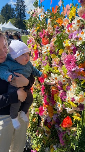 A mother holds her baby in front of a wall of flowers.
