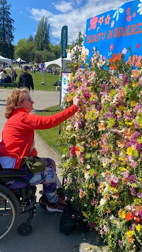 Woman in wheelchair adds a flower to a wall of flowers, under a sign that reads 'Beauty in Diversity'.