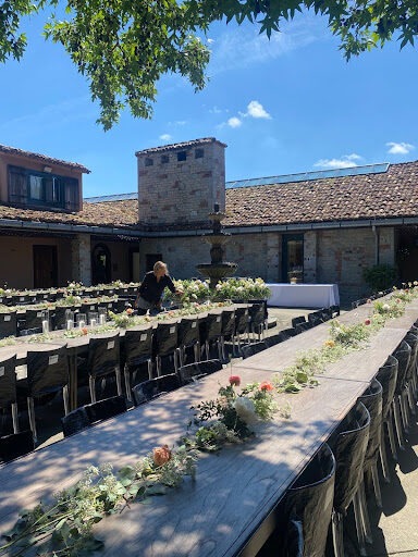 Flowers decorate two long tables outdoors in a courtyard.