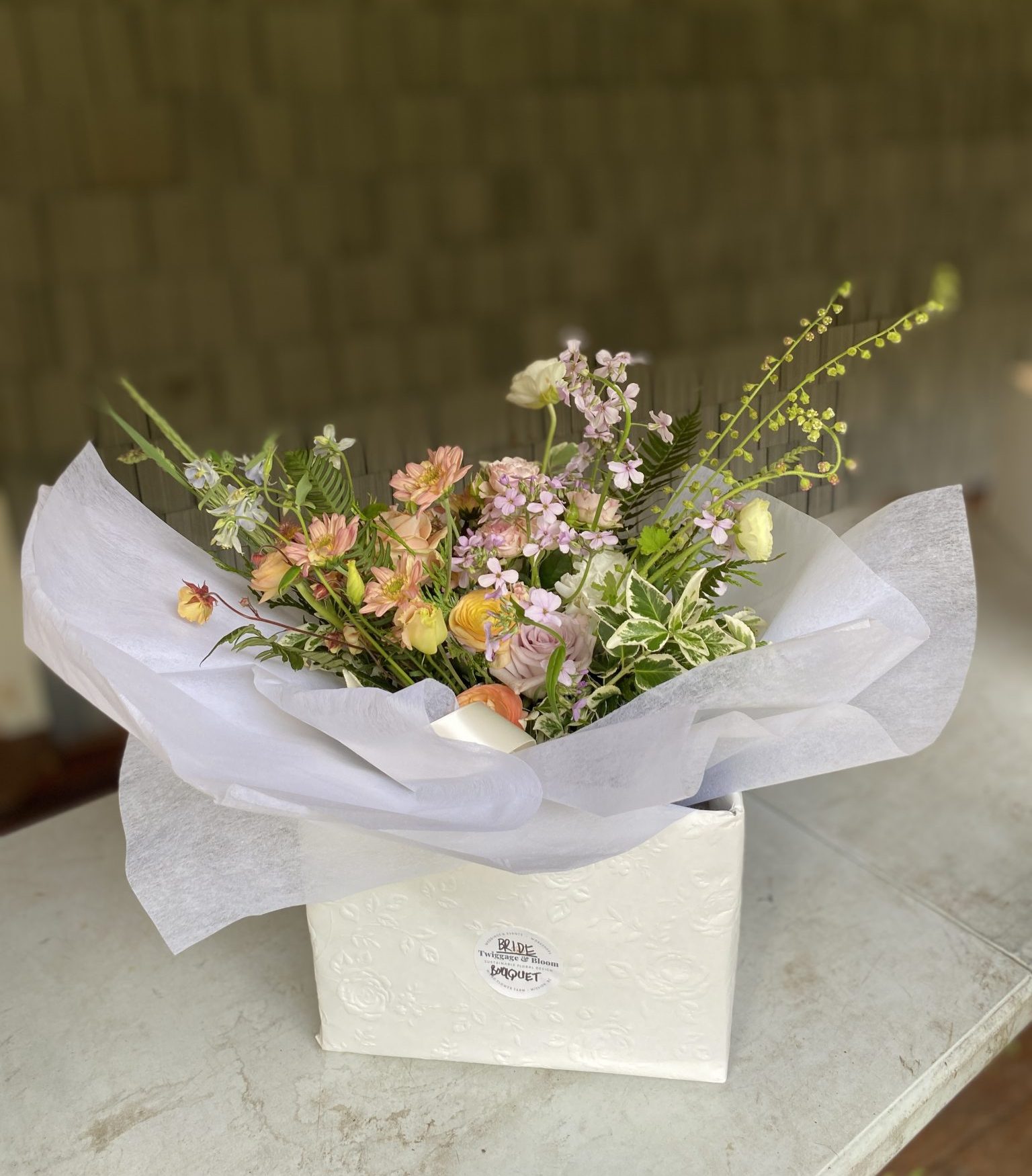 A bridal bouquet nestled in a white embossed gift box with white tissue paper, featuring peach, blush, and lavender blooms with airy greenery. The box is labeled "Bride — Twiggage & Bloom Bouquet."