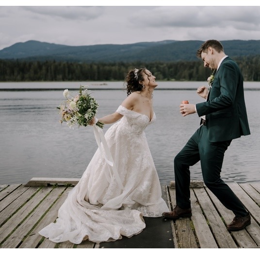 A laughing bride in an off-shoulder lace gown and groom in a dark green suit dance playfully on a wooden dock overlooking a lake with forested mountains in the background. The bride holds a lush wildflower bouquet by Twiggage and Bloom.