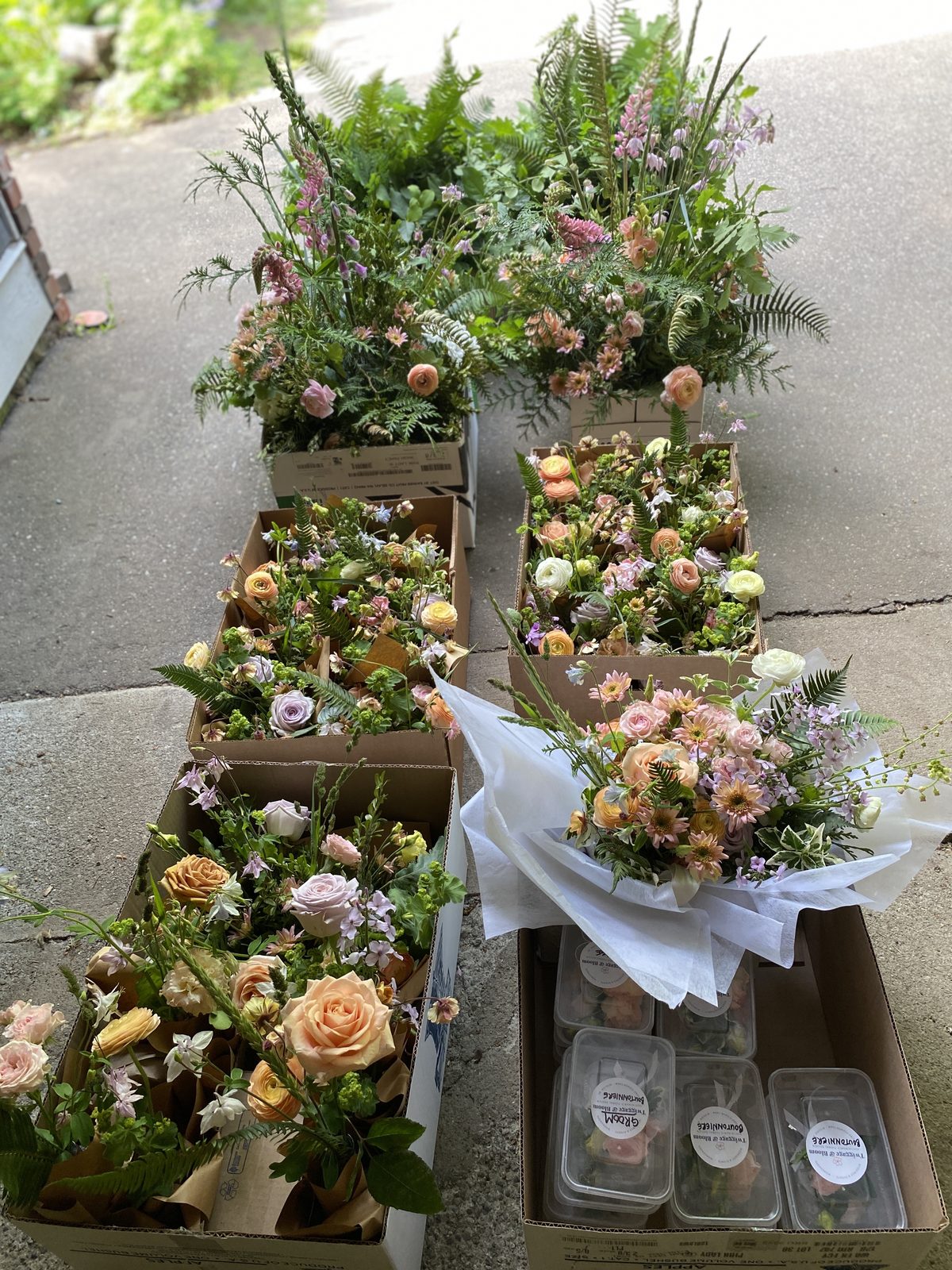 Multiple cardboard boxes spread out on a driveway, packed with wedding florals by Twiggage and Bloom, including centrepieces, bridesmaid bouquets, and a wrapped bridal bouquet in peach, blush, and lavender tones. Several labelled boutonnière containers for the groom and groomsmen are visible in the foreground.
