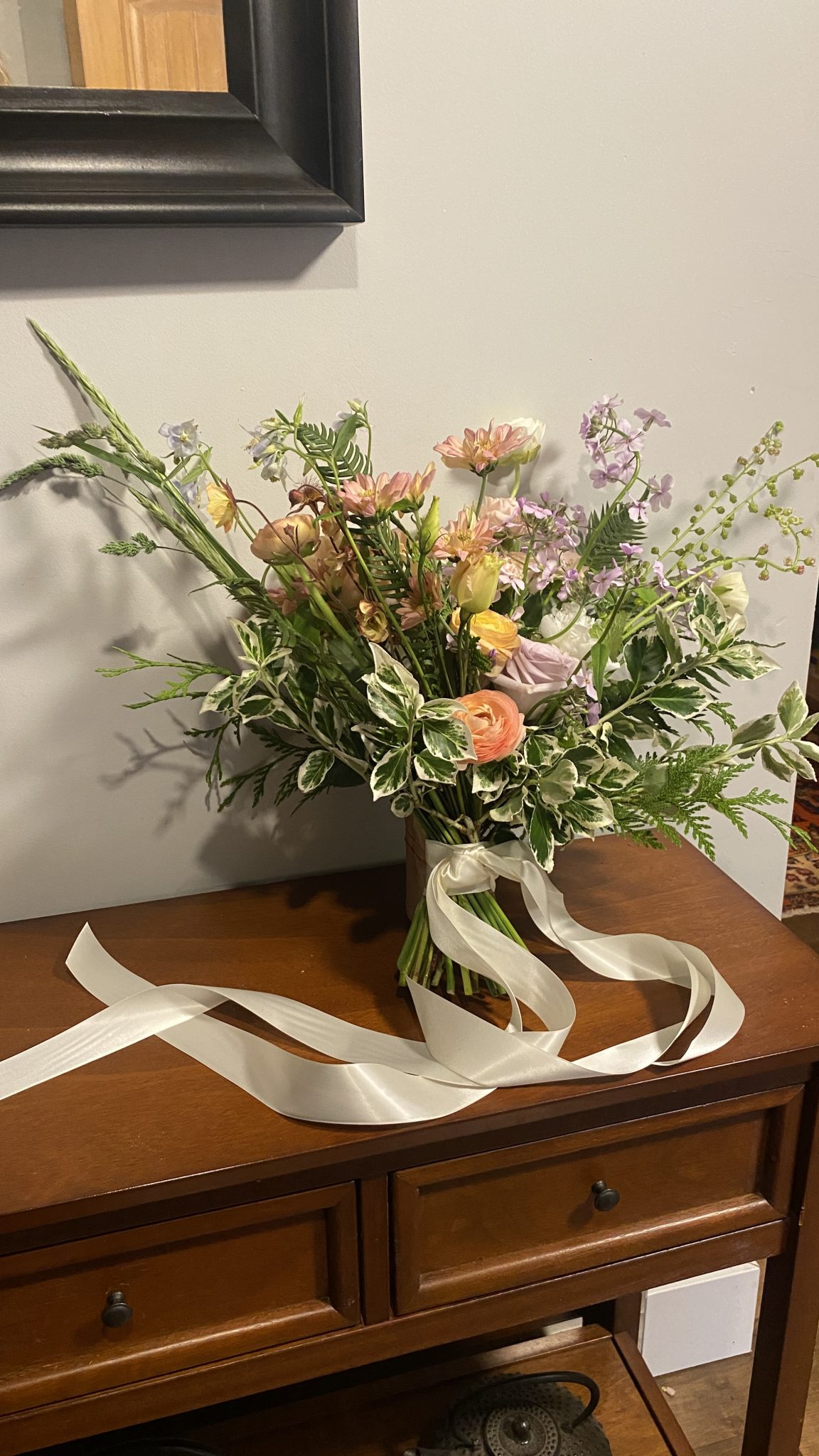 A loose, garden-style bridal bouquet resting on a wooden dresser, featuring peach ranunculus, lavender roses, blush dahlias, soft purple wildflowers, and variegated foliage, tied with long flowing white satin ribbon. Floral design by Twiggage and Bloom.